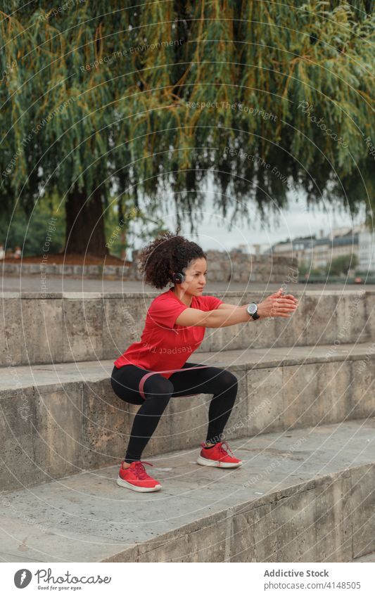 Ethnischer Sportler mit intelligenter Uhr, der mit einem elastischen Band im Freien hockt Sportlerin Kniebeuge Klebeband Training Übung intelligente Uhr Headset