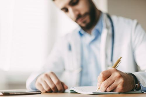 Freundlicher junger männlicher Arzt mit orientalischem Aussehen sitzt an einem Schreibtisch in seinem Büro und macht sich Notizen in einem Notizbuch, Nahaufnahme