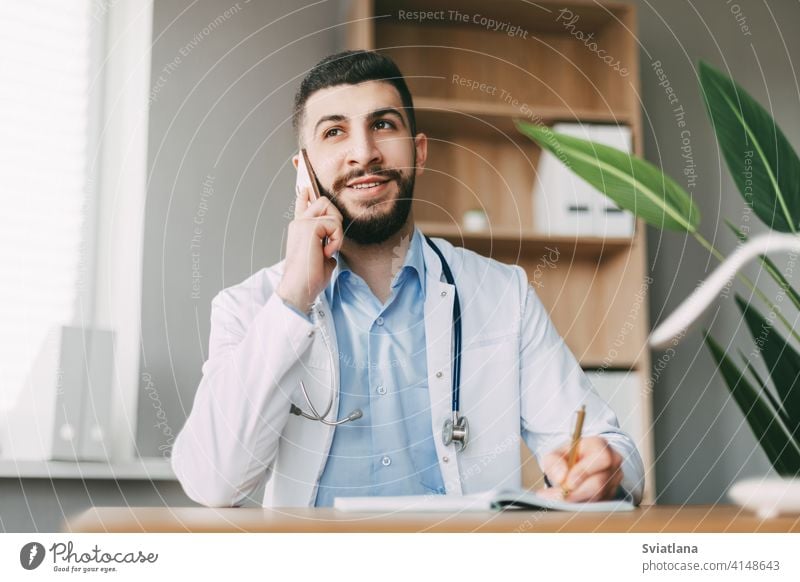 Ein junger Arzt mit orientalischem Aussehen telefoniert, sitzt am Tisch und macht sich Notizen in einem Notizbuch Medizin Mann Gesundheit medizinisch