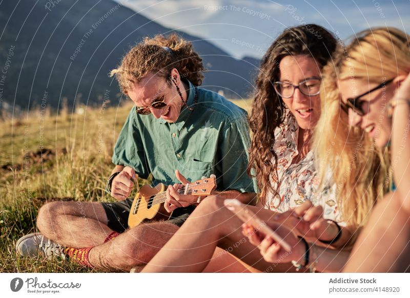Zufriedene Freunde, die gemeinsam in den Bergen entspannen spielen Ukulele sich[Akk] entspannen Zusammensein Berge u. Gebirge Smartphone Hipster unterhalten
