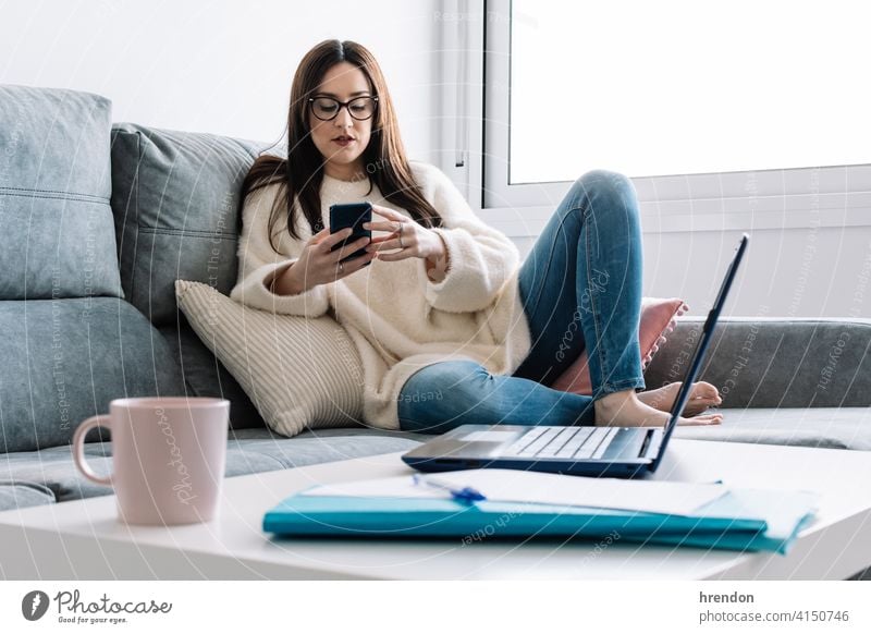 Frau im Gespräch mit Handy während der Arbeit mit Laptop sitzt auf einer Couch zu Hause im Innenbereich benutzend arbeiten Büro Job Notebook jung Glück Tippen