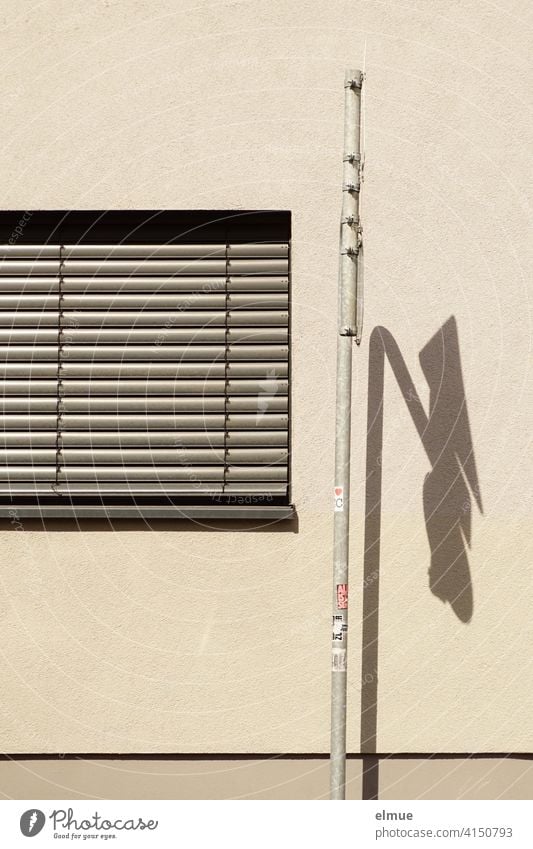 eine graue Jalousie verdeckt das Fenster neben einem Verkehrsschild mit Schattenwurf an der beigefarbenen Fassade / wohnen Fensterjalousie Fensterrollo Straße