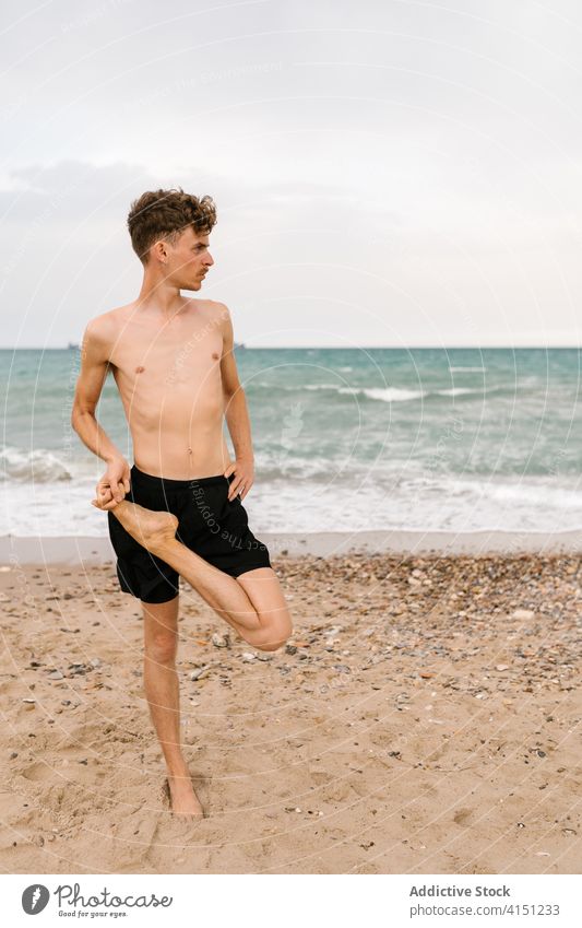 Flexibler junger Yogi am Strand stehend Mann Yoga üben Sand Halb-Lotus-Pose ardha padmasana Asana Gleichgewicht männlich beweglich schlank ohne Hemd