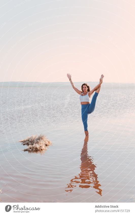Schlanke Frau dehnt sich in Yogapose am Seeufer üben Asana Pose stehen Split thiruvikramasana Gleichgewicht Wasser Windstille Harmonie Wellness Lifestyle
