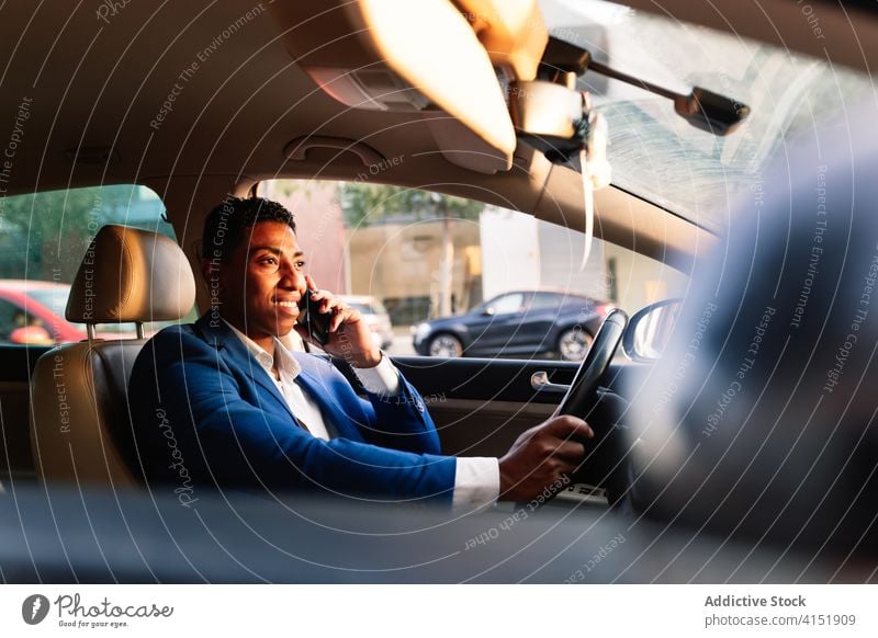 Lächelnder ethnischer Geschäftsmann, der im Auto mit seinem Smartphone spricht PKW Laufwerk reden Glück Kommunizieren positiv urban formal jung Mann