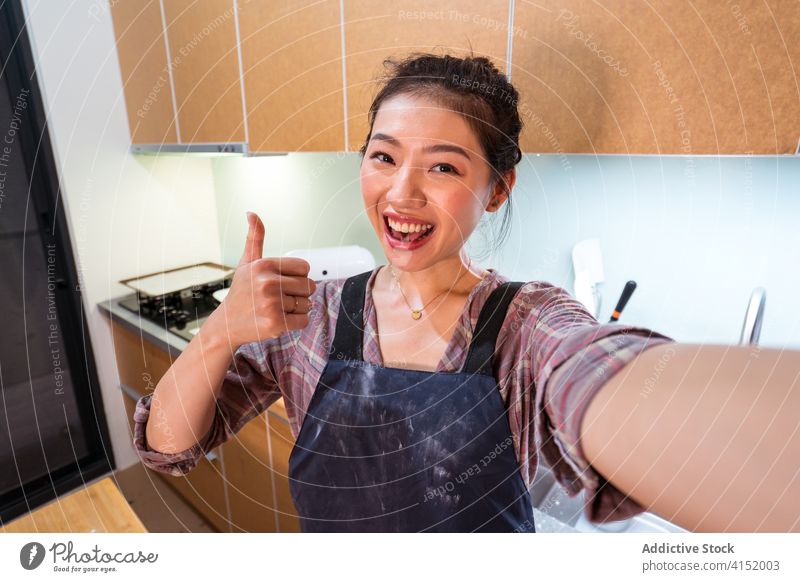 Lachender ethnischer Bäcker zeigt Daumen nach oben und macht ein Selfie Frau heimwärts Daumen hoch Glück zufrieden Küche heiter aufgeregt soziale Netzwerke