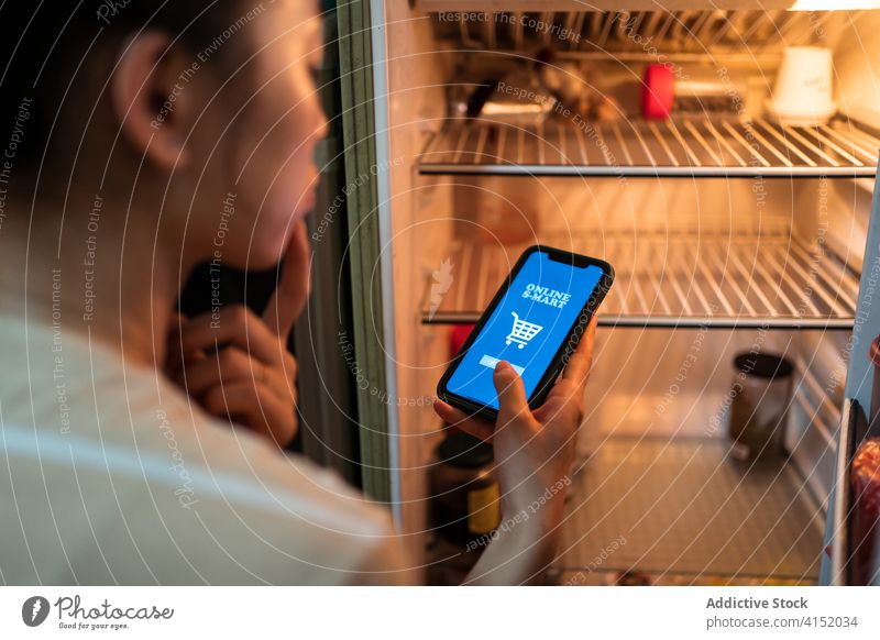 Frau mit Smartphone in der Nähe eines leeren Kühlschranks Orden Lebensmittel online App Werkstatt benutzend nachdenklich Apparatur Gerät Mobile Internet Telefon