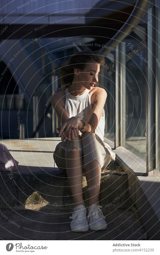 Entspannte junge Frau sitzt in der Nähe von Glaszaun in der Stadt besinnlich sich[Akk] entspannen Stil urban Gebäude nachdenklich Windstille verträumt modern