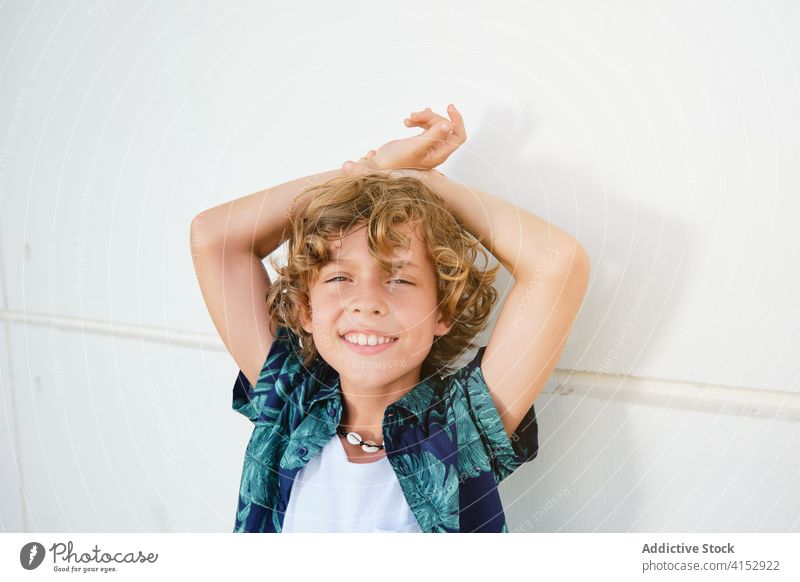 Junge in Sommerkleidung, der sich an eine Wand lehnt, während er der Kamera zugewandt ist vertikal Kinder selbstbewusst Arme posierend positiv Hemd Model