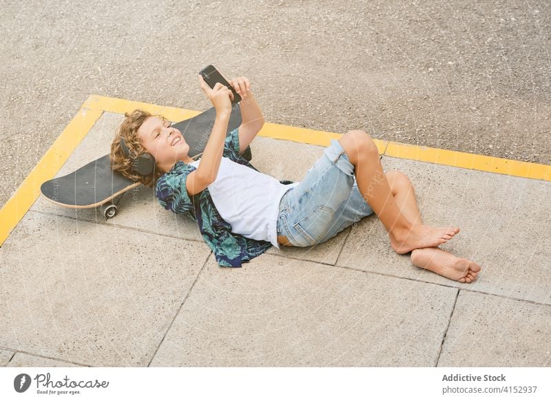 Lächelnder Junge, der mit einem Skateboard auf dem Boden liegt und mit seinem Handy Musik hört laut Jugendzeit zuhören Einsamkeit Gesang Stilrichtung Generation