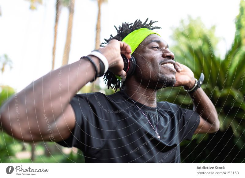 Nachdenklicher ethnischer Sportler, der mit seinen Kopfhörern Musik hört gestikulieren hören Athlet sportlich Gelassenheit Park Training Stil jung