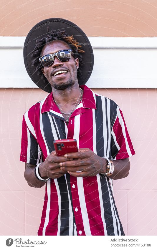 Fröhlicher stilvoller Mann mit Smartphone auf der Straße stehend Stil Mode benutzend trendy Glück heiter farbenfroh Sonnenbrille Streifen Hut Mobile Telefon