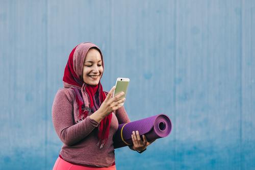 Lächelnde ethnische Sportlerin mit Smartphone in der Stadt benutzend Browsen Frau Hijab muslimisch Unterlage arabisch Großstadt Training Telefon Mobile Straße