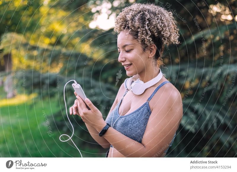 Fröhliche ethnische Sportlerin mit Smartphone im Park Frau sportlich benutzend ruhen heiter passen jung Browsen Talkrunde Kopfhörer Training Sommer Apparatur
