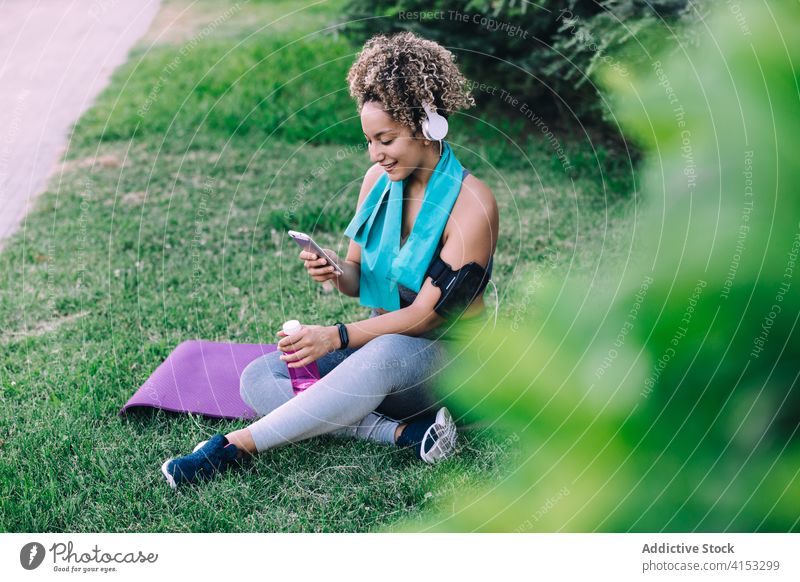 Sportliche Frau benutzt ihr Smartphone nach dem Training im Park sportlich benutzend ruhen heiter passen jung Browsen Kopfhörer Sommer Apparatur Fitness Gerät
