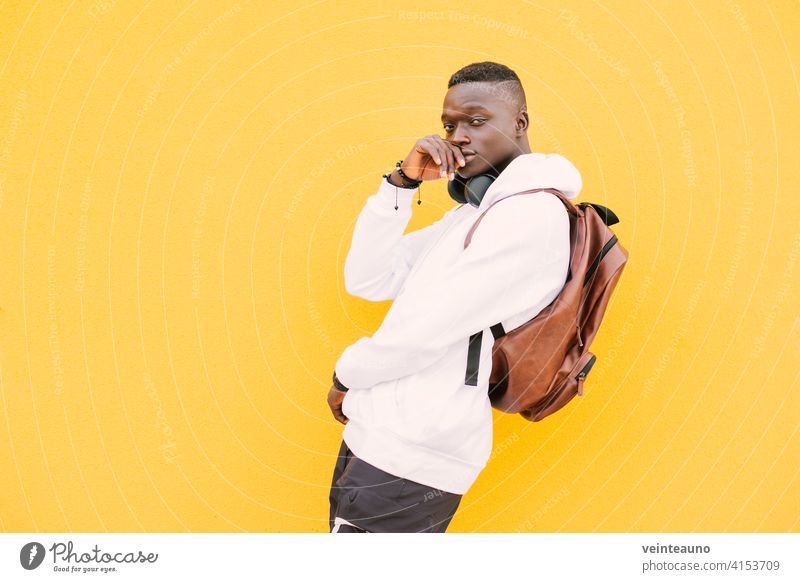 Junger afrikanischer amerikanischer schwarzer Mann gegen eine gelbe Wand, der ein weißes Sweatshirt und einen Rucksack trägt und Musik über drahtlose Kopfhörer hört, während er mit Haltung in die Kamera schaut