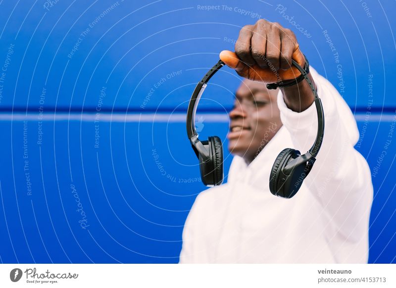Junger schwarzer Mann, der drahtlose Kopfhörer hält, während er ein weißes Sweatshirt trägt, vor einer blauen Wand, die selbstbewusst wegschaut. Selektiver Fokus