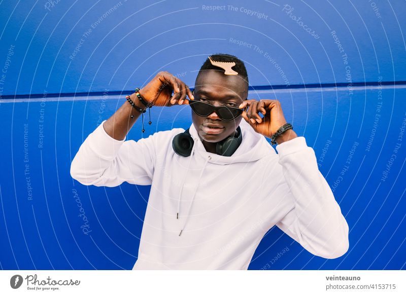 Junger afroamerikanischer schwarzer Mann mit weißem Sweatshirt, Sonnenbrille, Kamm und Kopfhörern an einer blauen Wand, der selbstbewusst über die Brille schaut