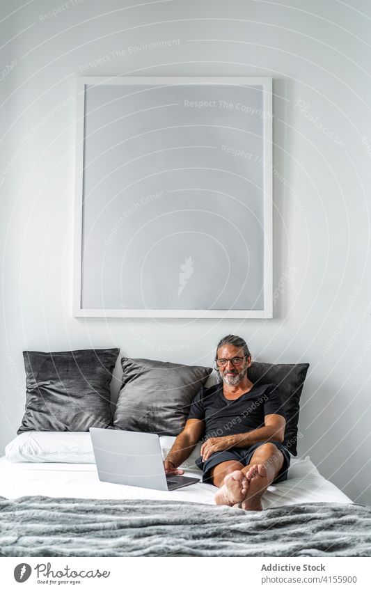 Älterer Mann auf dem Bett zu Hause freiberuflich abgelegen Arbeit Laptop Tippen reif Unternehmer benutzend männlich Browsen Internet Lächeln online Projekt