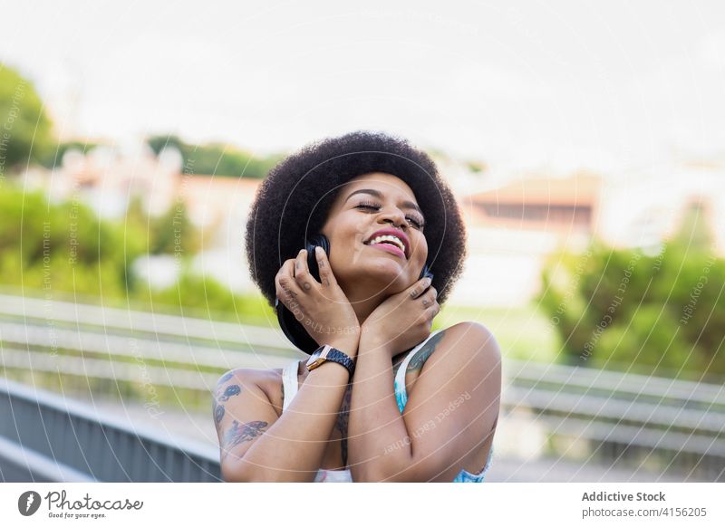 Fröhliche ethnische Frau beim Musikhören in der Stadt genießen Kopfhörer zuhören sorgenfrei Straße Sommer Drahtlos Freiheit schwarz Afroamerikaner Freude Gerät