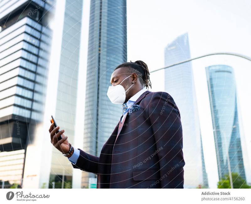 Seriöser ethnischer Geschäftsmann mit Maske in der Stadt Mundschutz Browsen behüten Coronavirus Smartphone zuschauend Stadtzentrum Großstadt Bestimmen Sie