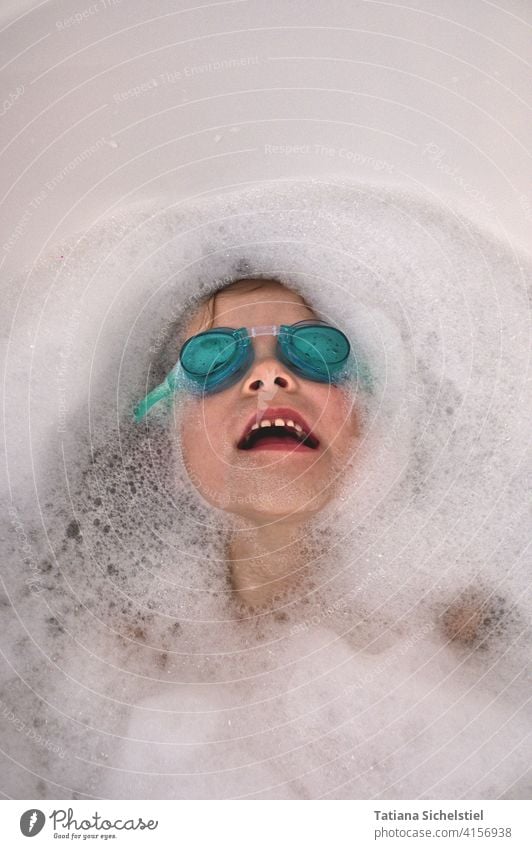 Kind liegt mit türkisfarbener Schwimmbrille in der Badewanne, nur das Gesicht ist zu sehen Baden Taucherbrille tauchen Schaum Wasser lachen Freude nass