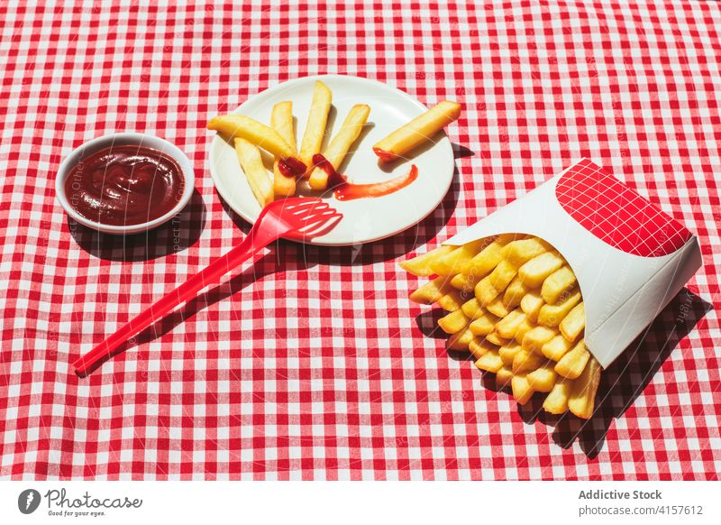 Pommes-Paket neben Teller mit in Ketchup getränkten Kartoffeln Amerikanische Kultur Gewürz Kartoffelchips ungesunde Ernährung roter Hintergrund weiß Fritten