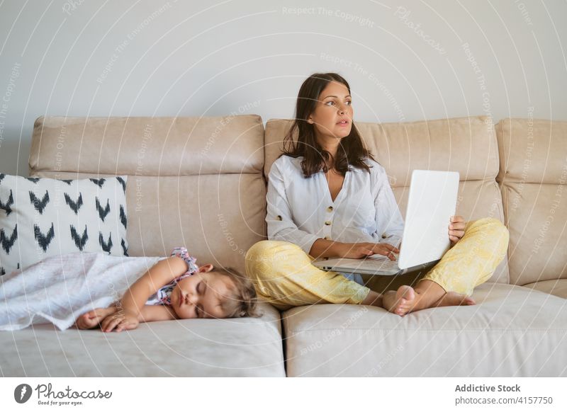 Abgelenkte Frau, die auf dem Sofa mit dem Laptop arbeitet, während ein Kleinkind auf einer Seite schläft Ruhe Mutterschaft Verantwortung Zärtlichkeit träumend