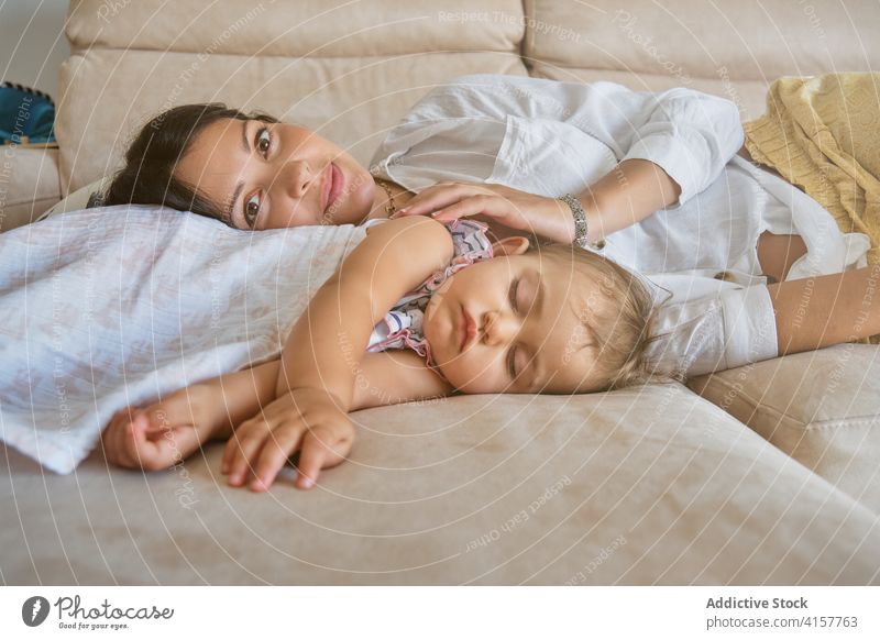 Frau schaut in die Kamera, während sie auf dem Sofa neben einem schlafenden Kleinkind liegt erschöpft streichelnd Vertrauen Zuneigung Umarmen Gefühl Säugling