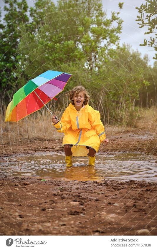 Kind im Regenmantel mit Regenschirm, zusammengekauert in einer Pfütze spielend und lachend Schuljunge Profil Dusche Einstellung Regenbogen lockig gestikulieren