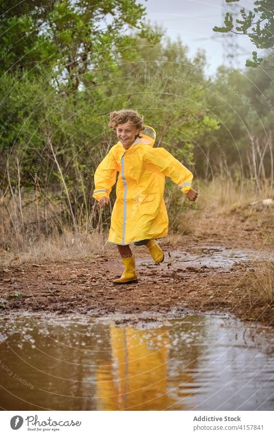 Junge in Regenmantel und Regenstiefeln, der auf einem Weg voller Pfützen läuft und spielt Lachen Schuljunge Dusche Einstellung lockig rennen Genuss