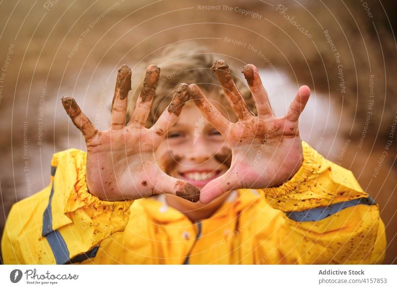 Selektiver Fokus auf die schlammigen Hände eines Kindes mit schlammigem Gesicht im Regenmantel im Freien Witz Stolz Säugling unordentlich geistig spielerisch