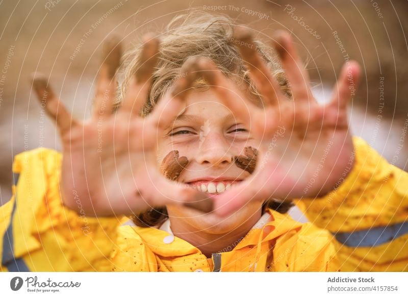 Selektiver Fokus auf das schlammige und glückliche Gesicht eines Kindes, das seine schmutzigen Hände im Regenmantel im Freien zeigt Witz spielerisch Säugling