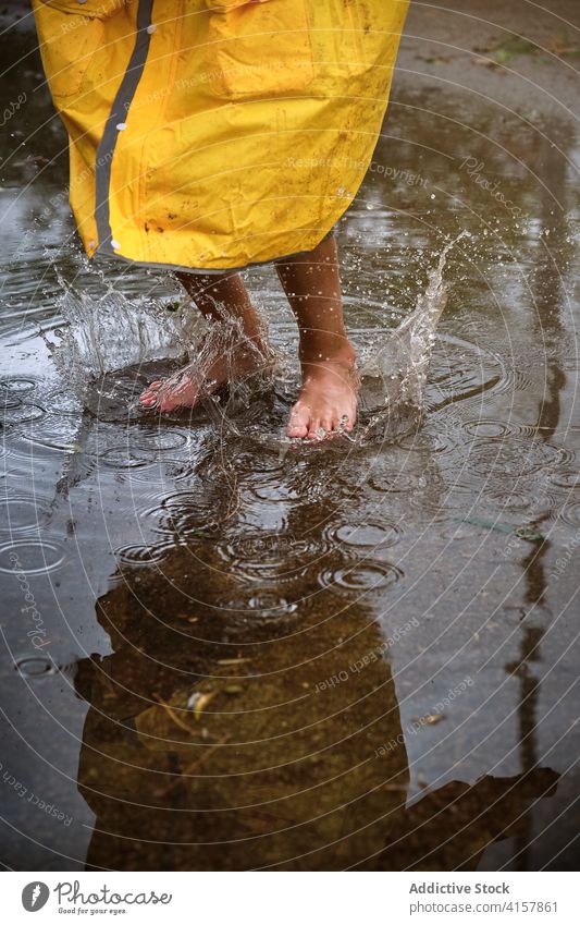 Barfüßiges Kind in einem gelben Regenmantel, das in eine Pfütze springt und Wasser verspritzt Barfuß springen platschen Genuss nass Fliege Wetter Freude spielen