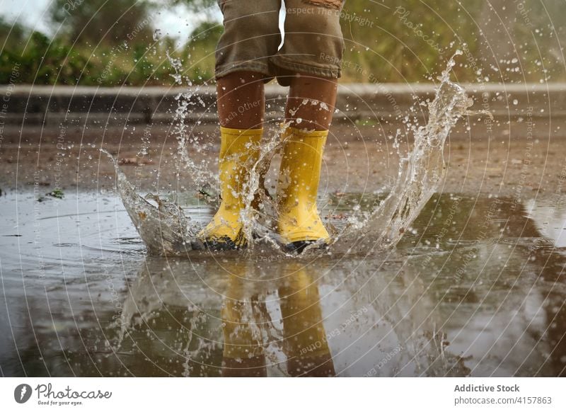 Detail von gelben Regenstiefeln, die auf eine Pfütze treffen und Wasser in einen Weg spritzen Witz Unschuld Lebensstile unordentlich springen spielerisch