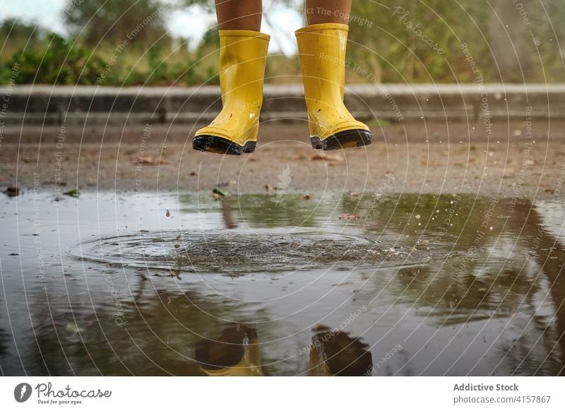 Beine eines Jungen mit gelben Regenstiefeln, der über eine Wasserpfütze auf einem Weg springt springen Witz Unschuld Lebensstile unordentlich spielerisch