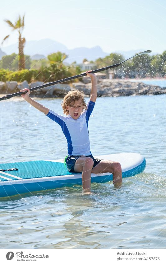 Junge mit lockigem Haar sitzt auf einem Paddel Surfbrett und spielt mit dem Paddel spritzt mit dem Wasser Vitalität spielen sitzen gestikulieren Waffen anheben