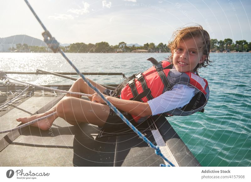 Junge mit Schwimmweste, der sich an den Rand eines Bootes lehnt und das Ruder und ein Seil im Meer hält fettarm Segeln Gewicht Meeresufer fahren Genuss Ferien
