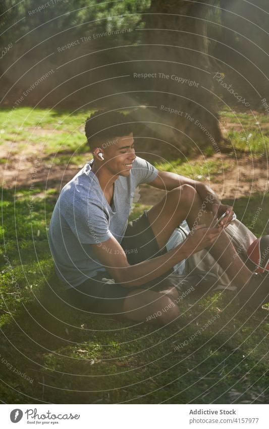 Schwarzer sportlicher Mann sitzt auf Rasen und benutzt Smartphone Sportler Basketball sich[Akk] entspannen Training Talkrunde Athlet Sommer männlich ethnisch