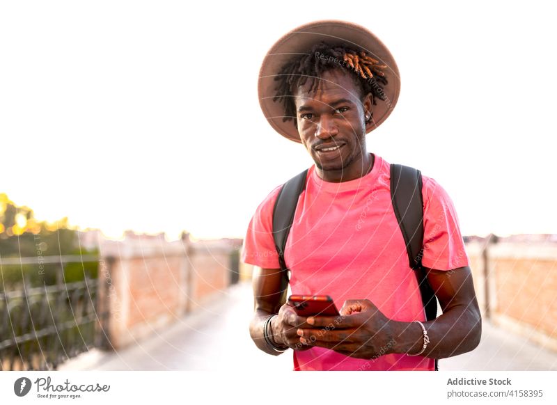Lächelnder ethnischer Mann auf einer Brücke im Sommer Reisender Smartphone Tourist gutaussehend benutzend Großstadt Sightseeing männlich schwarz Afroamerikaner