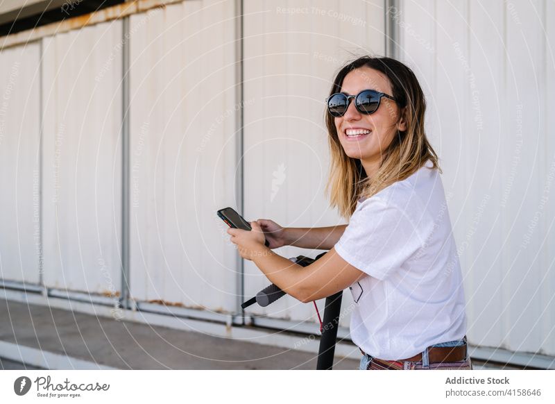 Fröhliche Frau mit Elektroroller und Smartphone benutzend heiter Mobile App Verkehr Mitfahrgelegenheit Glück urban Fahrzeug alternativ umweltfreundlich jung