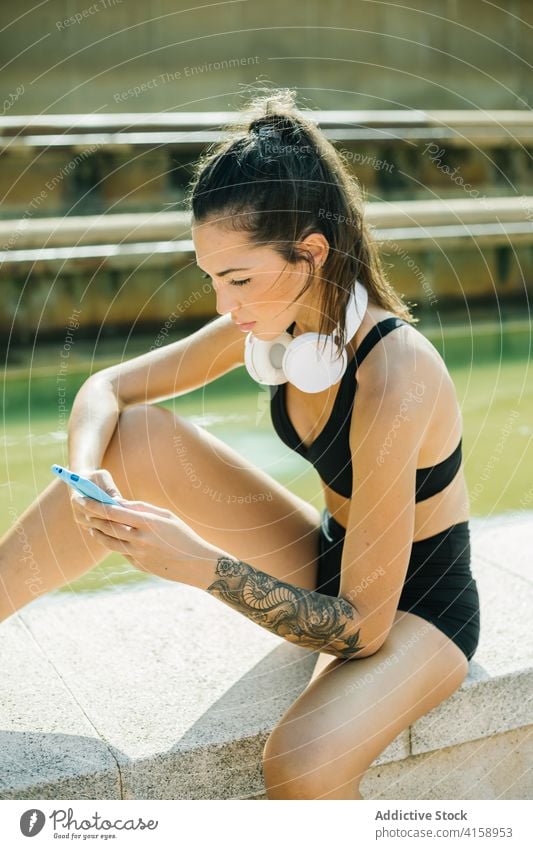 Sportliche Frau mit Smartphone und Kopfhörern ruht sich nach dem Training auf der Straße aus Sportlerin benutzend ruhen Browsen Apparatur Athlet Tattoo Mobile