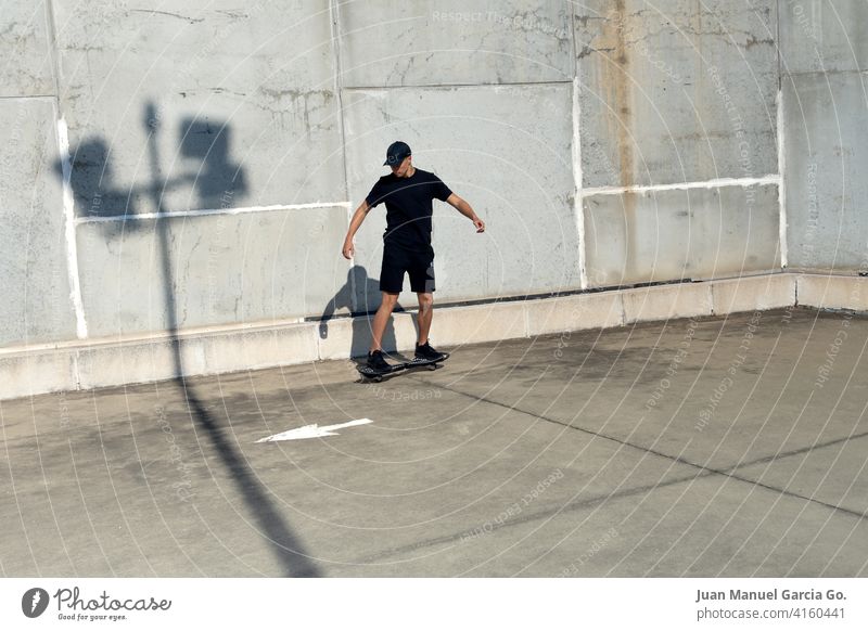 Schwarz gekleideter Teenager mit Mütze beim Skateboarden im Industriegebiet Parkplatz Verlassen abgestufter Haarschnitt solo Dachterrasse Ernsthaftigkeit