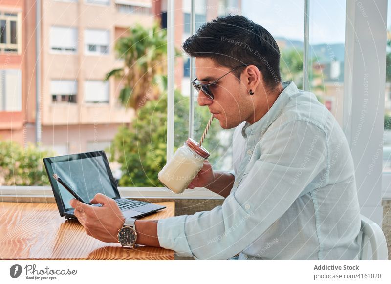 Konzentrierter junger Mann, der ein Getränk trinkt, während er aus der Ferne mit Laptop und Smartphone arbeitet trinken Milchshake benutzend Arbeit