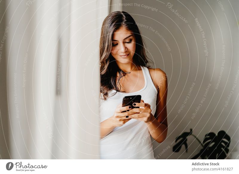 Junge Frau auf Smartphone stehen in der Nähe von Fenster am Morgen benutzend heimwärts Browsen Apparatur tausendjährig Lifestyle Schlafzimmer Gerät Internet