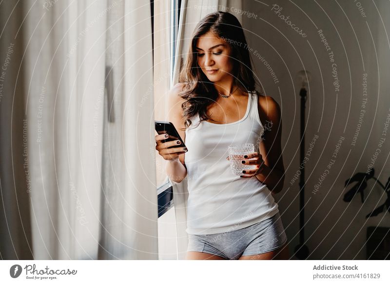 Junge Frau mit einem Glas Wasser und einem Smartphone am Fenster stehend am Morgen trinken benutzend heimwärts Browsen Apparatur tausendjährig Lifestyle