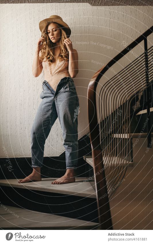 Stilvolle Frau steht auf einer Treppe in einem Haus lässig Hut Jeanshose Vorschein Outfit charmant sich[Akk] entspannen Treppenhaus hölzern modern stehen