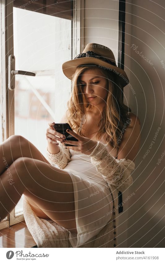 Stilvolle, verführerische Frau, die in der Nähe des Fensters mit ihrem Smartphone chattet plaudernd stylisch Bekleidung sinnlich feminin benutzend Apparatur