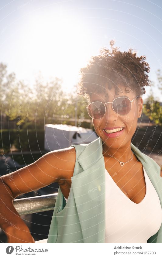 Stilvolle ethnische Frau mit Sonnenbrille in der Nähe von Zaun stehen trendy Glück Sommer Afro-Look Lächeln heiter jung Afroamerikaner schwarz positiv
