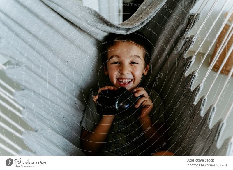 Aufgeregter Junge mit Fotokamera in der Hängematte zu Hause Fotoapparat Lachen aufgeregt Fotograf Hobby expressiv ruhen Freude Zeitgenosse Gerät Fotografie